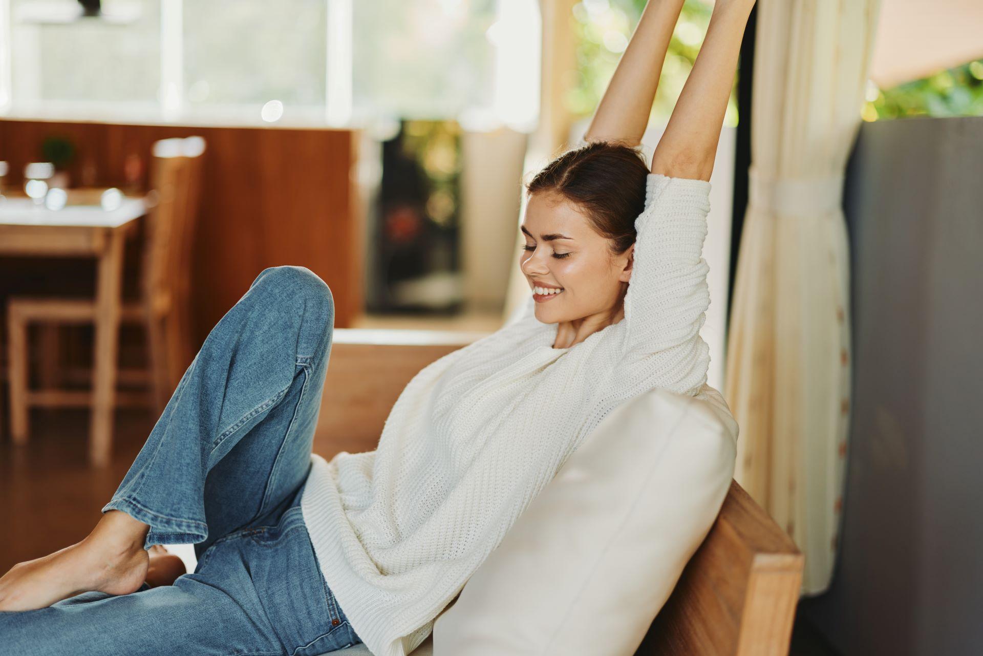 Frau sitzt zu Hause auf einer gemütlichen Couch und trägt einen warmen Strickpullover. Ihre natürliche Schönheit strahlt durch ihr freudiges Lächeln und ihren entspannten Gesichtsausdruck, während sie einen Moment der Zufriedenheit und des Glücks genießt
