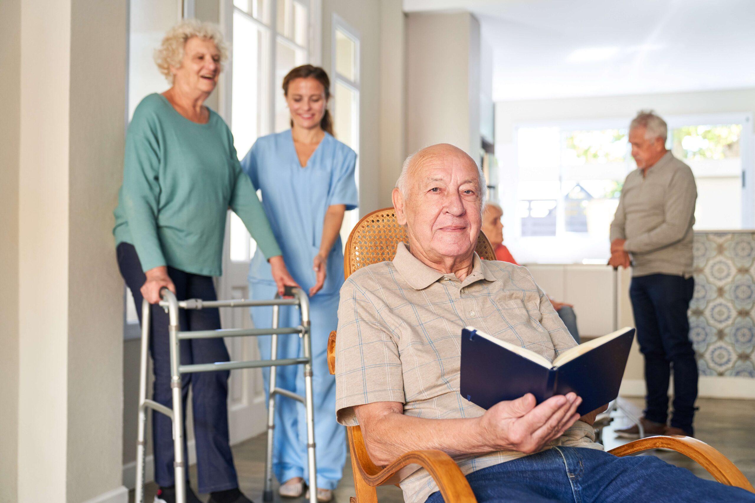 Ein älterer Mann sitzt in einem Stuhl und liest ein Buch, während eine Betreuungsassistentin in blauer Uniform einer älteren Frau mit Gehwagen hilft. Weitere Senioren sind im Hintergrund zu sehen.