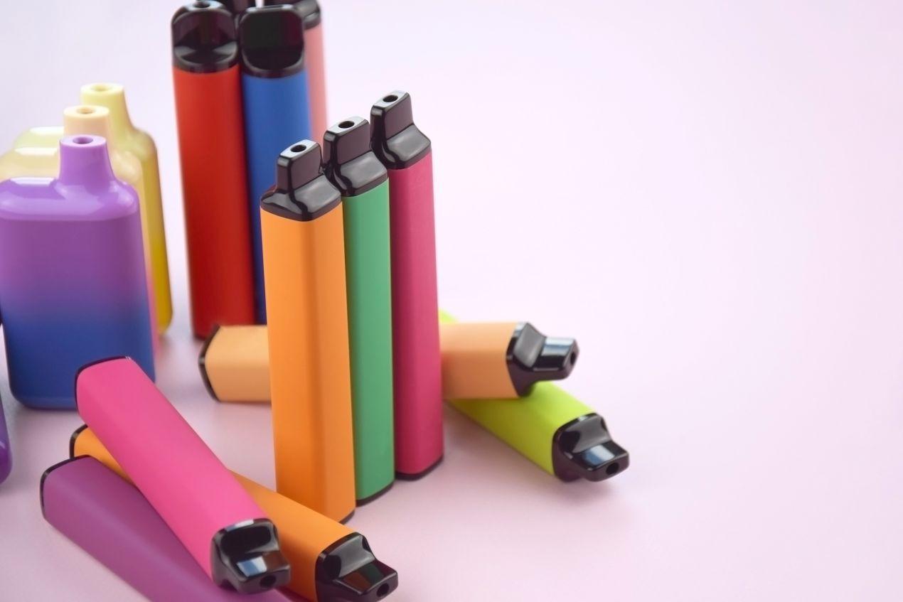 Mehrere bunte E-Zigaretten in leuchtenden Farben stehen und liegen auf einem hellrosa Hintergrund – modernes Design, wie es bei der Elfa Master beliebt ist